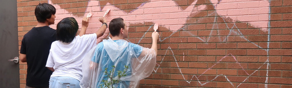 Three youth students painting a mural on the brick wall located on the side of the library